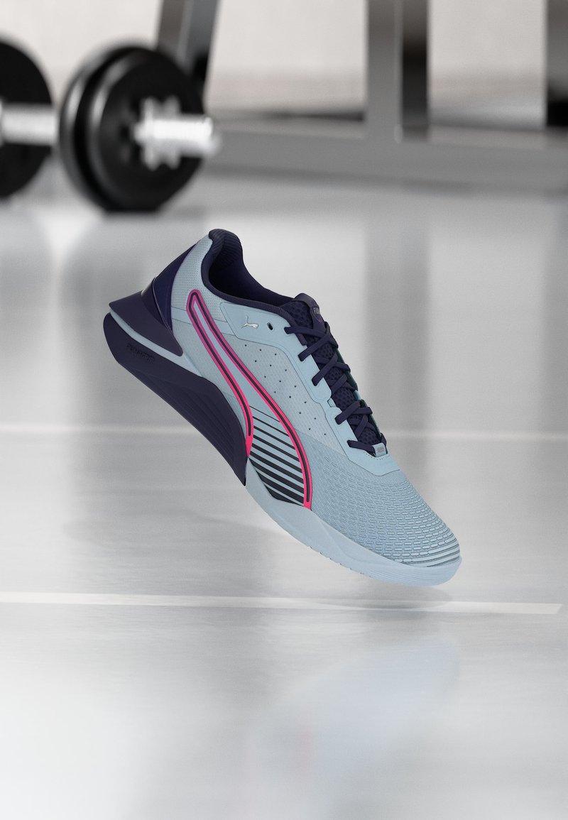 Jasnoniebieski i granatowy but sportowy z różowym paskiem, na tle podłogi na siłowni z rozmytym sprzętem do podnoszenia ciężarów.