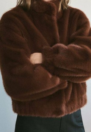 Personne portant une veste en fausse fourrure marron à manches longues, les bras croisés, associée à un pantalon noir sur un fond uni.