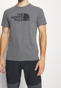 Camiseta de algodón gris con mangas cortas, que presenta un logo negro "THE NORTH FACE" en el frente. Cuello redondo simple y corte estándar.