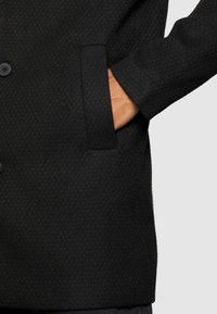 Černý vlněný kabát s texturovaným povrchem, vybavený postranní kapsou a zapínáním na tři knoflíky. Design je přiléhavý s upraveným střihem.