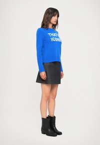 Blå strikkegenser med hvit tekst, kombinert med et svart denim miniskjørt og svarte ankelstøvletter. Enkle og moderne designelementer.