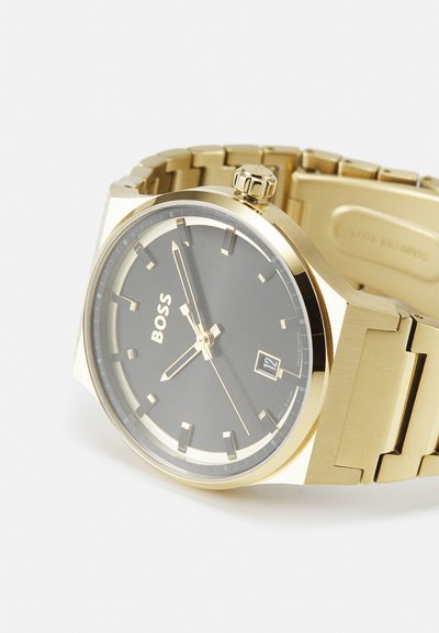 Montre-bracelet en métal doré avec un cadran gris. Dispose d'un affichage de la date à 6 heures et de trois aiguilles. Les maillons du bracelet sont polis et mats.