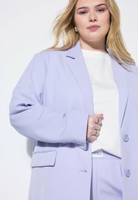Blazer de color púrpura claro, hecho de una tela suave, con un solo botón, solapas muestradas y un bolsillo en el pecho, usado sobre una blusa blanca.
