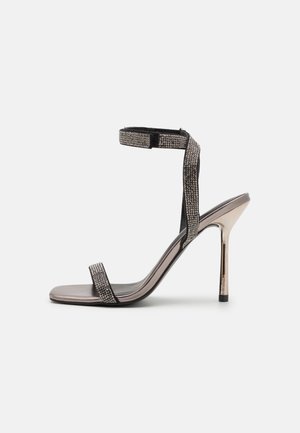 Sandaal met hoge hak en dunne metalen naaldhak, open teen en enkelbandje versierd met kleine strass-steentjes.
