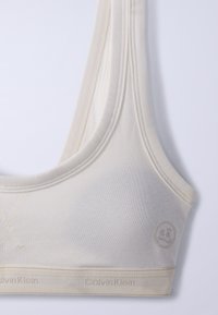 Αθλητικό σουτιέν Calvin Klein σε απαλό ύφασμα κρεμ χρώματος με φαρδιές τιράντες και λωρίδα με λογότυπο σε ανοιχτό φόντο.