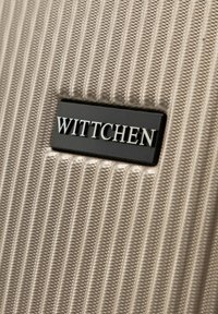 Zbliżenie na beżową, teksturowaną powierzchnię walizki z prostokątną czarną metką, na której widnieje nazwa marki "WITTCHEN" w srebrnych literach.