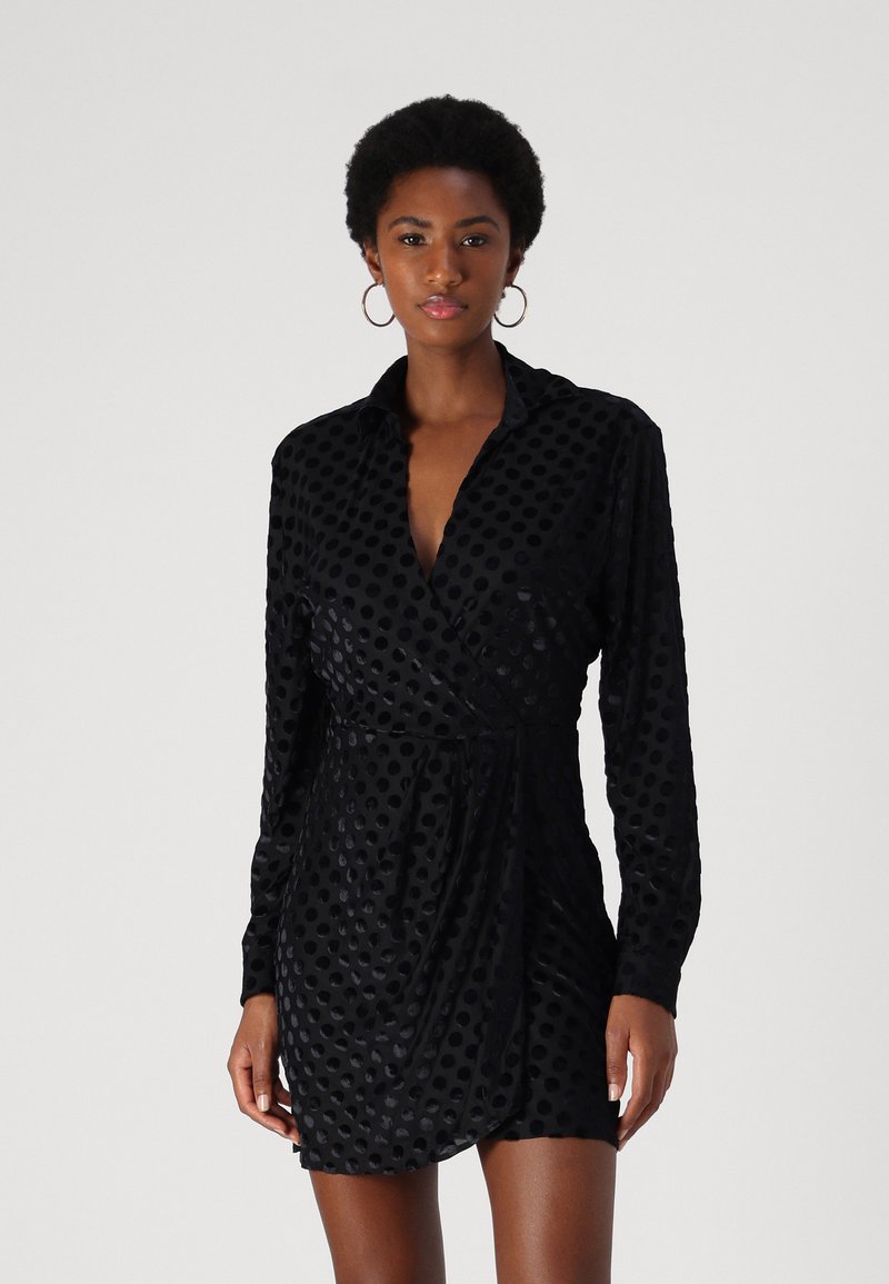 Robe noire portefeuille avec un motif à pois texturé, manches longues et col classique. Matière douce et lisse avec une silhouette ajustée.