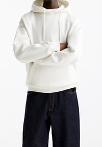 Persoon in een witte hoodie met een voorzak, met gekruiste armen, gecombineerd met donkerblauwe jeans tegen een eenvoudige witte achtergrond.