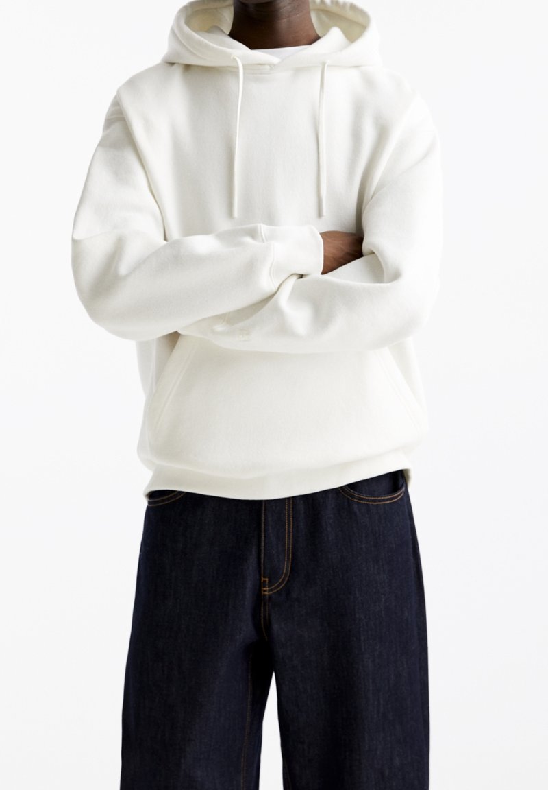 Persoon in een witte hoodie met een voorzak, met gekruiste armen, gecombineerd met donkerblauwe jeans tegen een eenvoudige witte achtergrond.