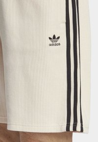 Vita ribbade shorts med svart tre-stripes-design, med en Adidas-logotyp på vänster ben. Materialet verkar mjukt och texturerat.