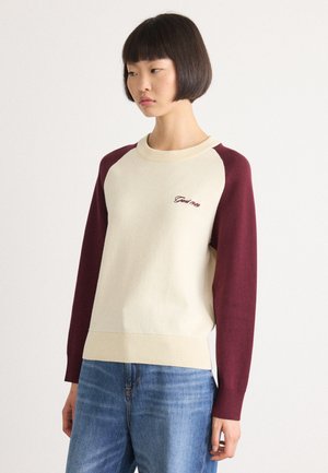 Woman with short bob haircut wearing a cream sweater with burgundy sleeves and light blue jeans, standing against a plain background.