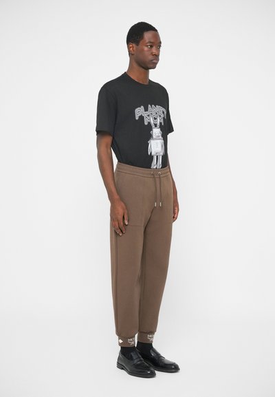 Man staat in een zwart graphic T-shirt met "PLANET MCM" en een robotontwerp, bruine joggers met koord, zwarte sokken en zwarte instappers.