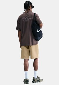 Camisa marrón de manga corta con el gráfico "Air's Everywhere"; pantalones cortos beige con el logo blanco de Nike; mochila negra de Nike; zapatillas oliva; calcetines blancos.