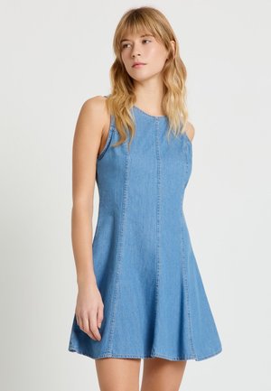 Donna con capelli biondi che indossa un vestito di jeans azzurro senza maniche, in piedi contro uno sfondo chiaro e uniforme.