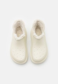Kremowe buty wsuwane z futrzanym wykończeniem wnętrza, charakteryzujące się gładką powierzchnią i okrągłymi otworami wentylacyjnymi w okolicy palców. Płaska gumowa podeszwa.