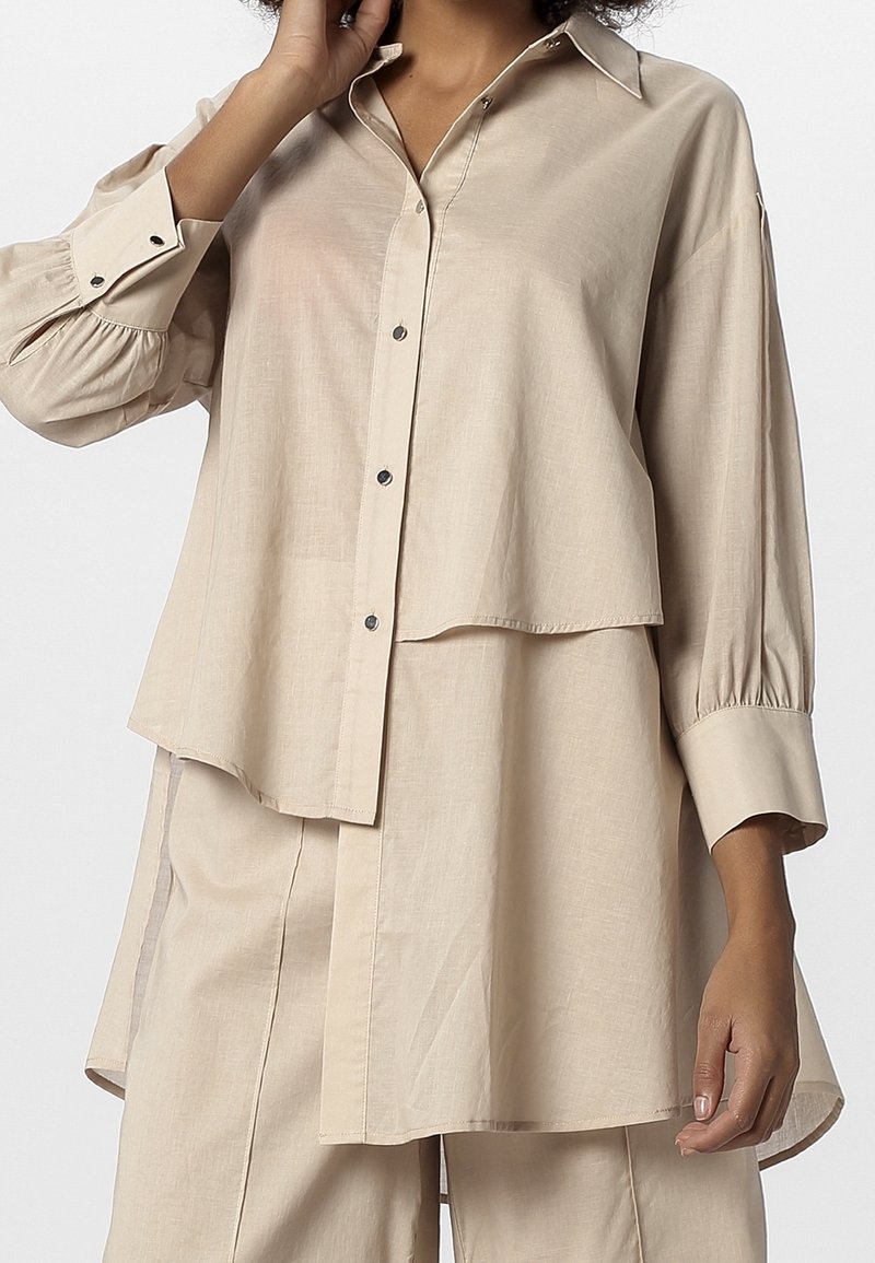 Femme portant une chemise boutonnée beige surdimensionnée à couches avec manches longues froncées et un pantalon beige assorti, sur fond uni.