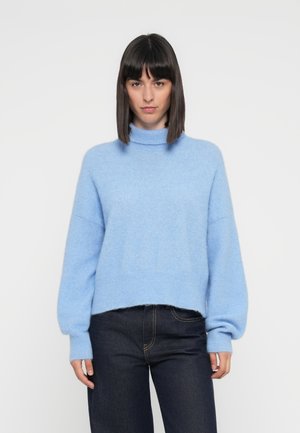 Lys blå, fluffy strikkegenser med høy hals, nedfalne skuldre og ballongsleeves, sammen med mørke jeans med synlige sømmer.