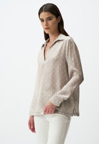Blusa beige a maniche lunghe con scollatura a V, realizzata in tessuto leggero, con pois bianchi e una texture leggermente trasparente.