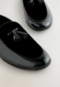 Schwarze Halbschuhe aus Lackleder mit einem Obermaterial aus Samt. Sie verfügen über eine runde Zehenpartie und dekorative schwarze Quasten auf der Vorderseite.
