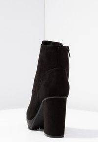 Botín negro de ante con un tacón alto y grueso, que cuenta con un cierre lateral y una textura suave, destacando un diseño elegante y moderno.