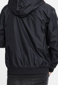 Veste noire légère avec capuche, poignets et ourlet élastiques. Matière en nylon texturée avec une finition lisse. Design simple et fonctionnel.