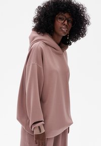 Overmaat roze hoodie van zacht materiaal met een plush binnenkant. Heeft een brede halslijn, ribgebreide mouwen en zichtbare stiksels.