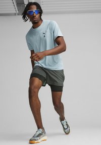 Světlomodré sportovní tričko, černé logo, v kombinaci s tmavě zelenými šortkami. Model nosí reflexní modré sluneční brýle a vzorované běžecké boty.