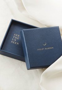 Marineblaue Box mit strukturiertem Finish. Hochwertig eingeprägter Text in Gold lautet "DIE UNSICHTBARE SEHEN" und "VIOLET HAMDEN." Der Deckel der Box ist leicht geöffnet und gibt den Blick ins Innere frei.