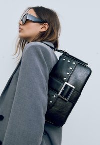 Sort læderrygsæk med sølvfarvede nitter og en rektangulær spænde. Har et struktureret design og justerbar strop, båret over en grå blazer.