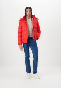 Rød puffjakke med lynlås og hætte, parret med en beige bluse og blå jeans. Jakken har vandret syninger og en blank overflade.