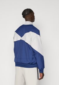 Giacca sportiva blu e bianca con un design a blocchi di colore geometrico. Caratterizzata da polsini a costine, colletto alto e vestibilità rilassata.
