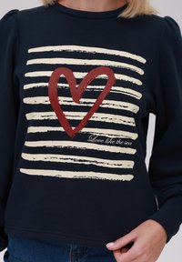 Sweatshirt marine avec des rayures blanches et un graphique en forme de cœur rouge. Le texte "Aime comme la mer" est présent sous le design. Tissu doux et texturé.