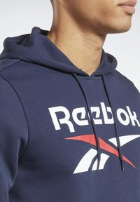 Mørkeblå hættetrøje med snore, der har et stort hvidt "Reebok" logo og et rødt diagonalt stribet design på brystet.