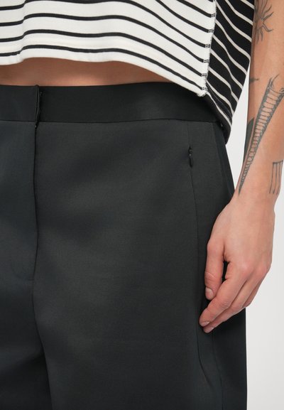 Pantaloni negri cu textură netedă, având un croi slim și buzunar lateral. Sub talie se ghicește un top cu dungi orizontale.