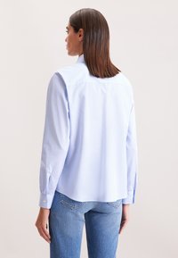 Donna con capelli castani lisci indossa una camicia a righe azzurra e jeans blu, in piedi di spalle contro uno sfondo neutro.