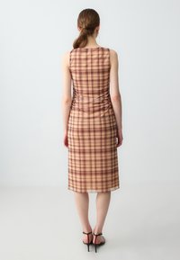 Karierter Kleid in Beige und Burgunder, figurbetonte Silhouette, knielang, mit seitlichen Rüschendetails und glatter Textur. Hochhackige Sandalen.
