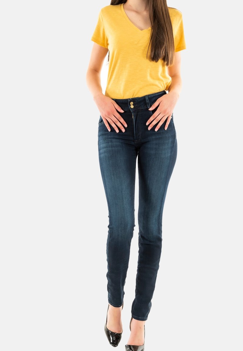 T-shirt jaune à col en V en tissu doux, associé à un jean skinny bleu foncé à taille haute, fermeture à bouton sur le devant et design épuré.