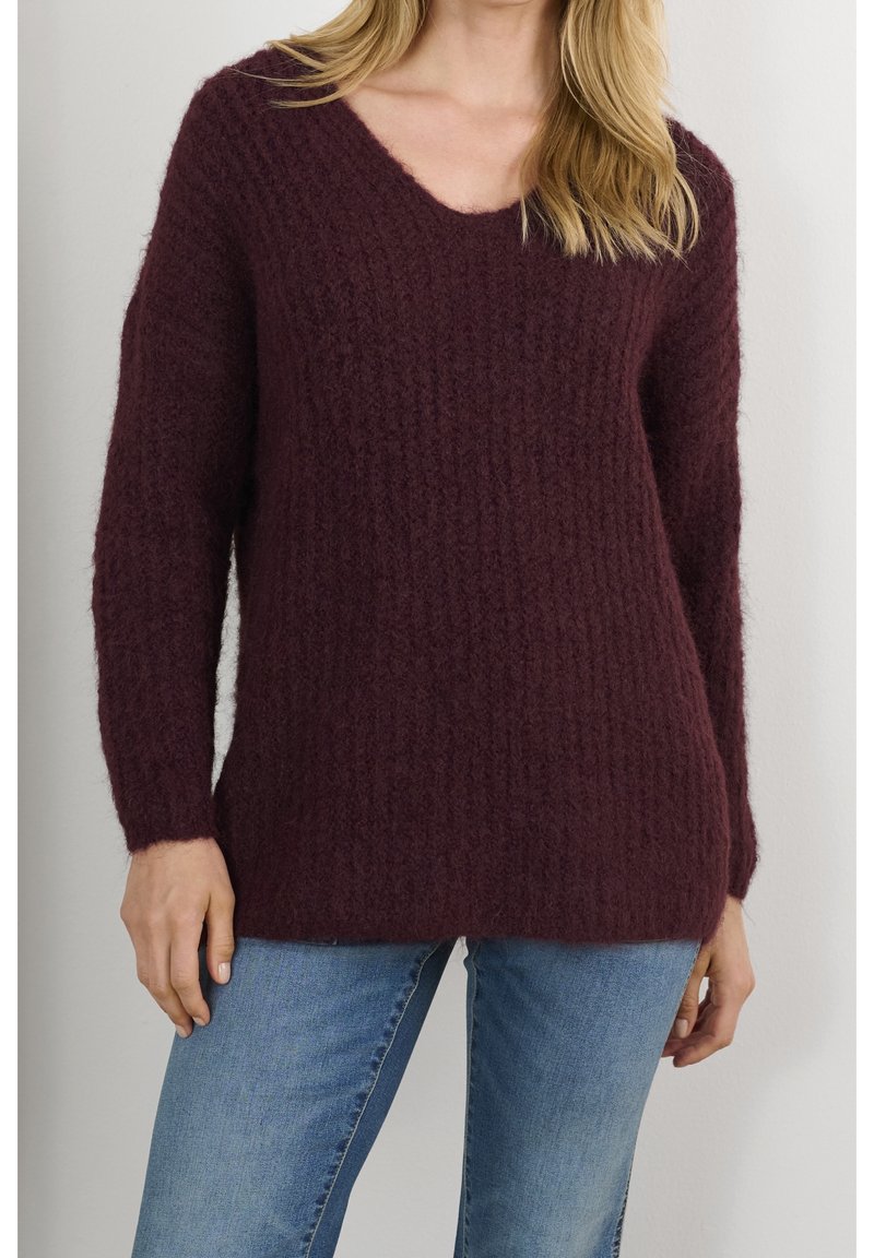 Femme portant un pull texturé à col en V violet foncé et un jean bleu clair, debout devant un fond clair uni.