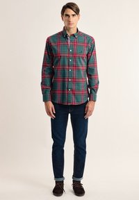 Camisa de cuadros verde y roja con botones, de mangas largas, con un collar estándar y un bolsillo en el pecho, combinada con jeans de mezclilla oscuros.