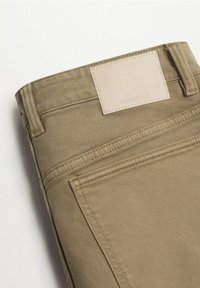 Pantalons en coton beige avec une texture lisse, présentant des coutures traditionnelles, une patch en cuir sur la taille et des poches arrière.