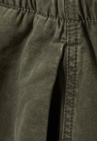 Olive green fabric with a textured surface, featuring an elastic waistband and visible stitching along the seams and pleats.