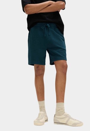 Mand iført mørkegrønne shorts med snor, beige sokker og beige sneakers stående med armene krydset.