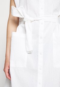 Vestido branco riscado, sem mangas, com um cinto na cintura, apresentando uma frente de botões e dois bolsos frontais, feito de tecido leve.