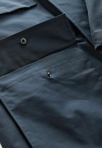 Tejido azul marino con dos bolsillos que presentan cierres a presión negros. Textura suave con detalles de costura visibles.
