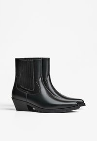 Par črnih usnjenih gležnjarjev s koničastimi prsti, nizkimi zloženimi petami in elastičnimi stranskimi paneli na beli ozadju.