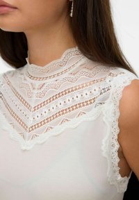 Mujer vestida con una blusa sin mangas blanca con intrincados detalles de encaje en el cuello alto y los hombros, y un arete de perla colgante.