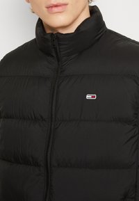 Uomo che indossa una giacca a vento nera trapuntata con colletto alto e piccolo logo ricamato sul petto, zip chiusa.