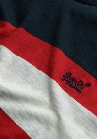 Close-up of fabric with horizontal stripes in navy, red, and white, featuring embroidered "SuperDry" logo in navy thread on the red stripe.