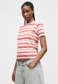 Γυναίκα με πλεξούδες, που φοράει ροζ και κόκκινη ριγέ κοντομάνικη μπλούζα και ανοιχτό μπλε τζιν, στέκεται με το ένα χέρι στην τσέπη.