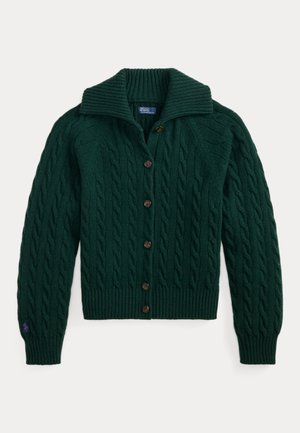 Femme aux cheveux tressés portant un cardigan en maille torsadée vert foncé, un jean bleu et des mocassins marron, debout devant un fond beige uni.